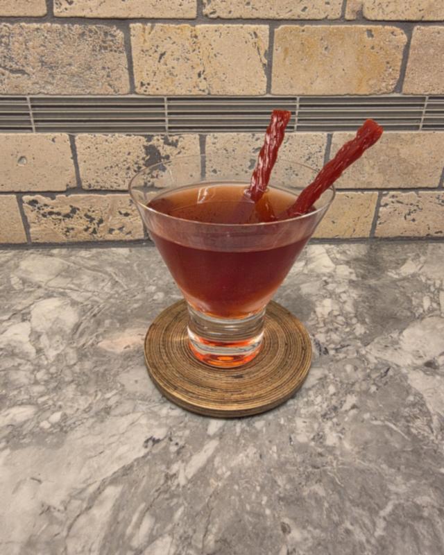 A deep cranberry red Vampire's Kiss in a stemless martini glass with two red licorice twists, on a wooden coaster in front of a stone tile wall
