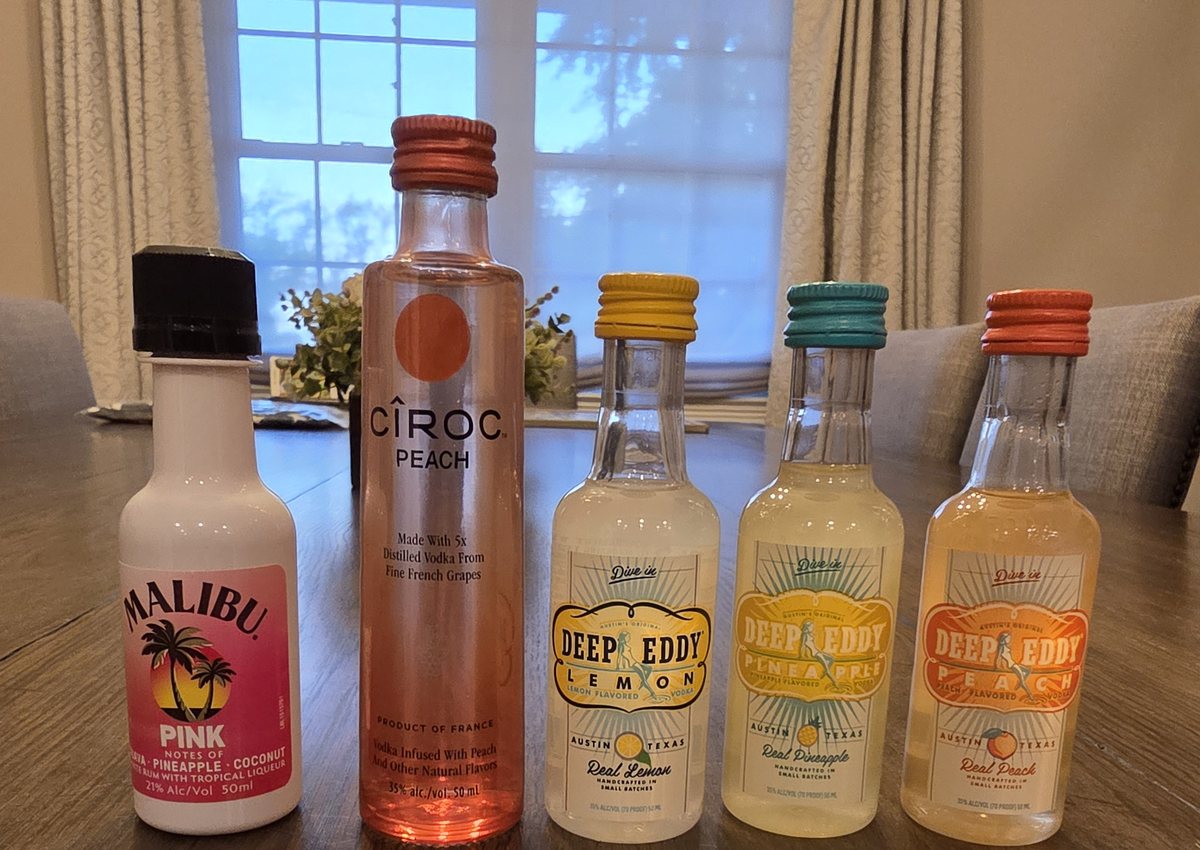 Five flavored spirit mini bottles, Deep Eddy Lemon, Pineapple and Peach vodka, Ciroc Peach, and Malibu Pink, lined up on a dining table